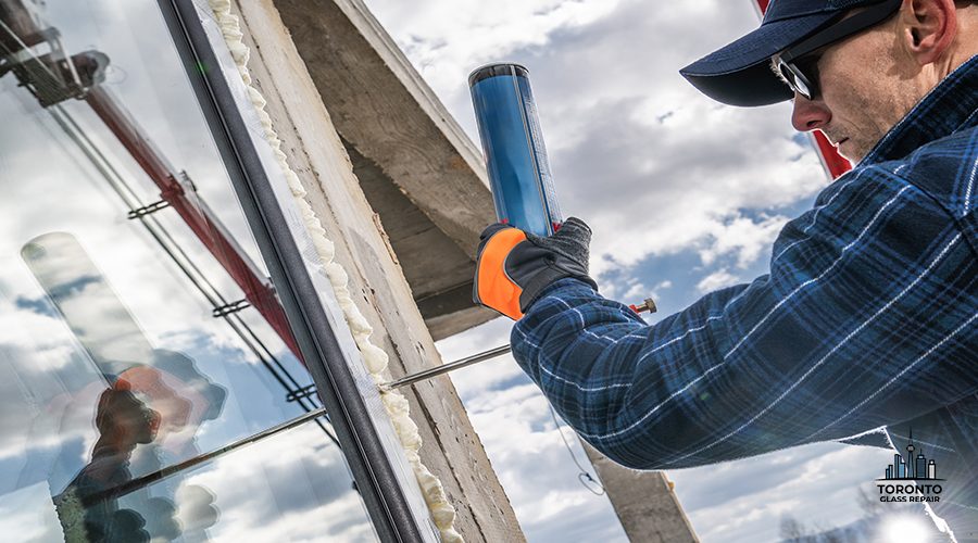 A construction worker applies sealant to install glass in a building while using a caulking gun. The cloudy sky serves as a backdrop, highlighting the ongoing construction activity during the day.